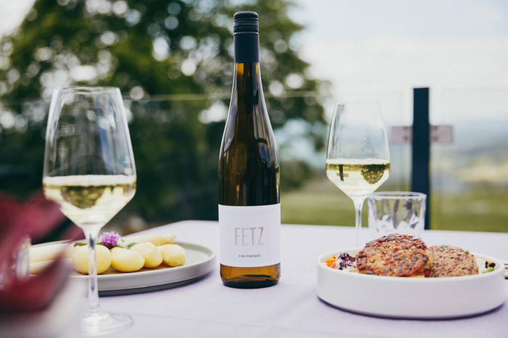 Ein gedeckter Tisch mit FETZ Weinflasche, Gläsern, Kartoffeln und vegetarischen Burgern im Freien.
